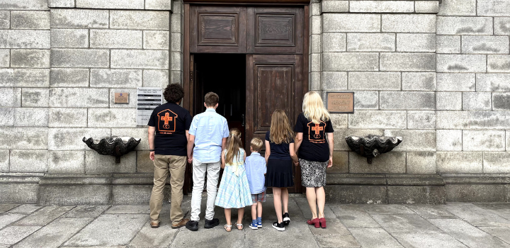 A family with mother and father with four children facing towards the door of a church. The mother and father are wearing Domestic Church t-shirts 