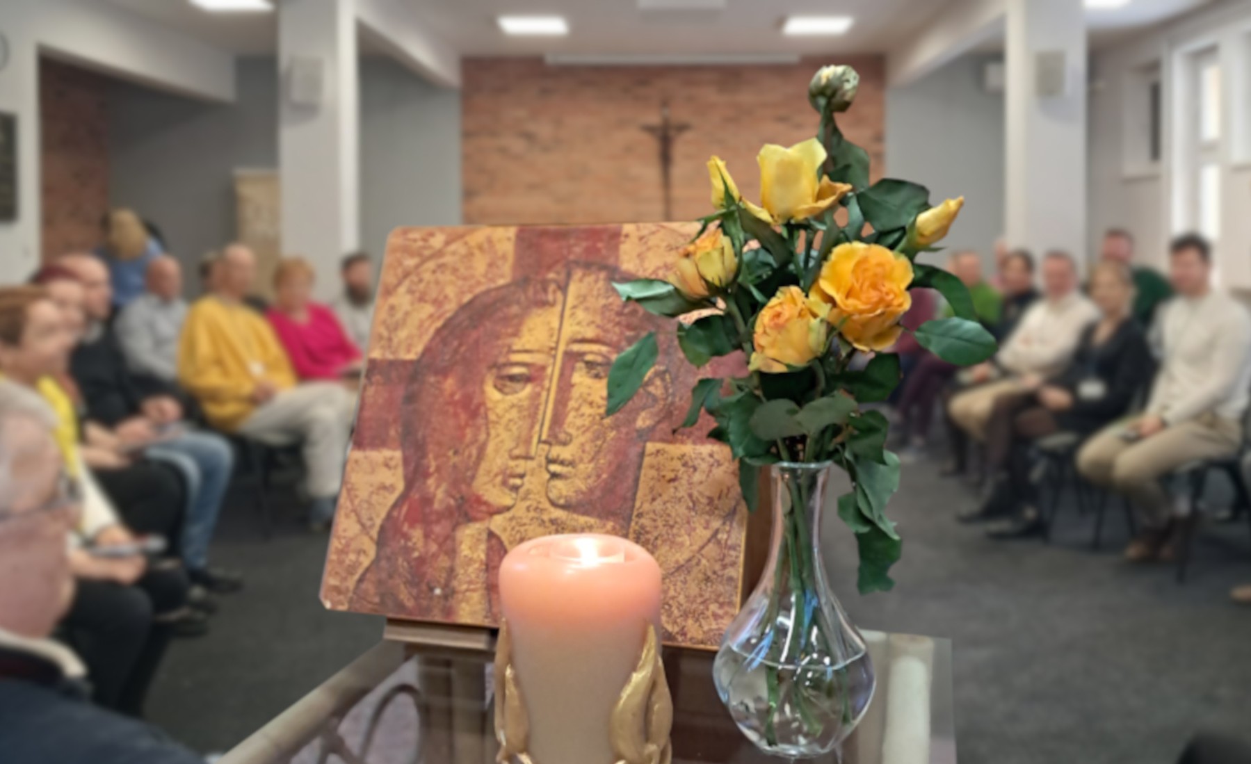 Table in a chapel with people gathered around. The table has an icon, candle and a vase of flowers