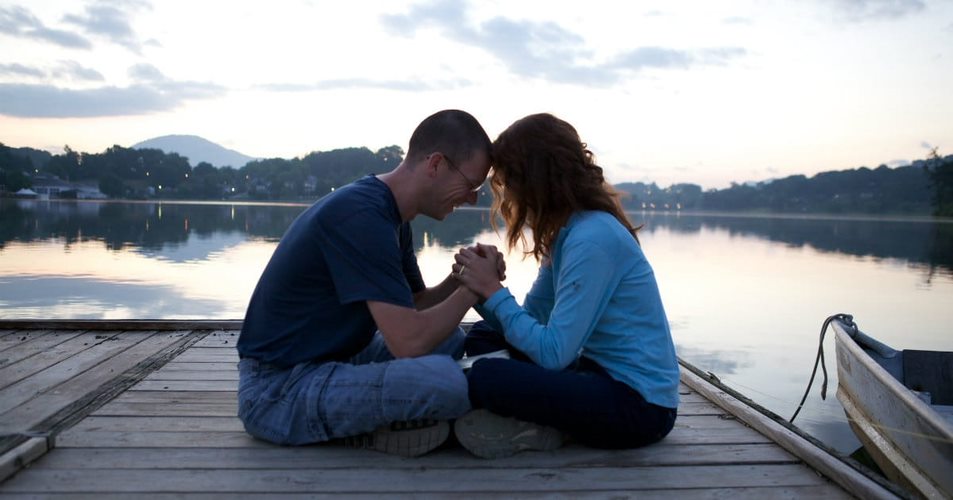 Picture of a couple sitting cross-legged, holding hands between them and touching foreheads. They are sitting on a wooden dock beside a lake